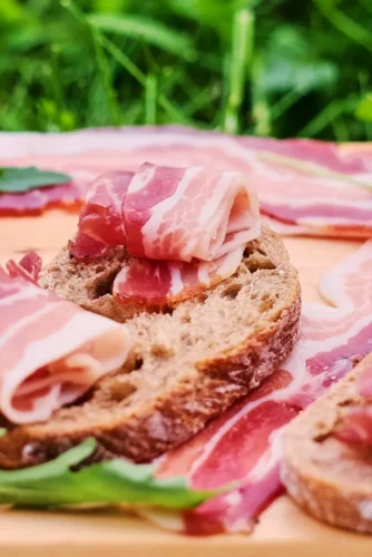 Tostas de pan integral con lonchas de panceta sobre una tabla de madera, acompañadas de hojas de rúcula y tiras de panceta en el fondo. Ideal para aperitivos o picnics.