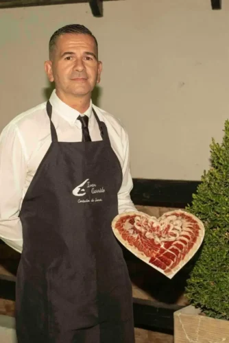 Hombre con delantal sosteniendo un plato en forma de corazón con embutidos, junto a un arbusto decorativo en un ambiente elegante.