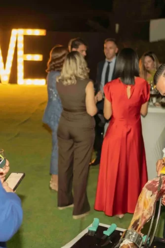 Celebración al aire libre con un grupo de personas disfrutando de una fiesta, mientras un camarero corta jamón en primer plano. Al fondo, una gran decoración iluminada con la palabra "LOVE". La escena refleja un ambiente festivo y elegante.